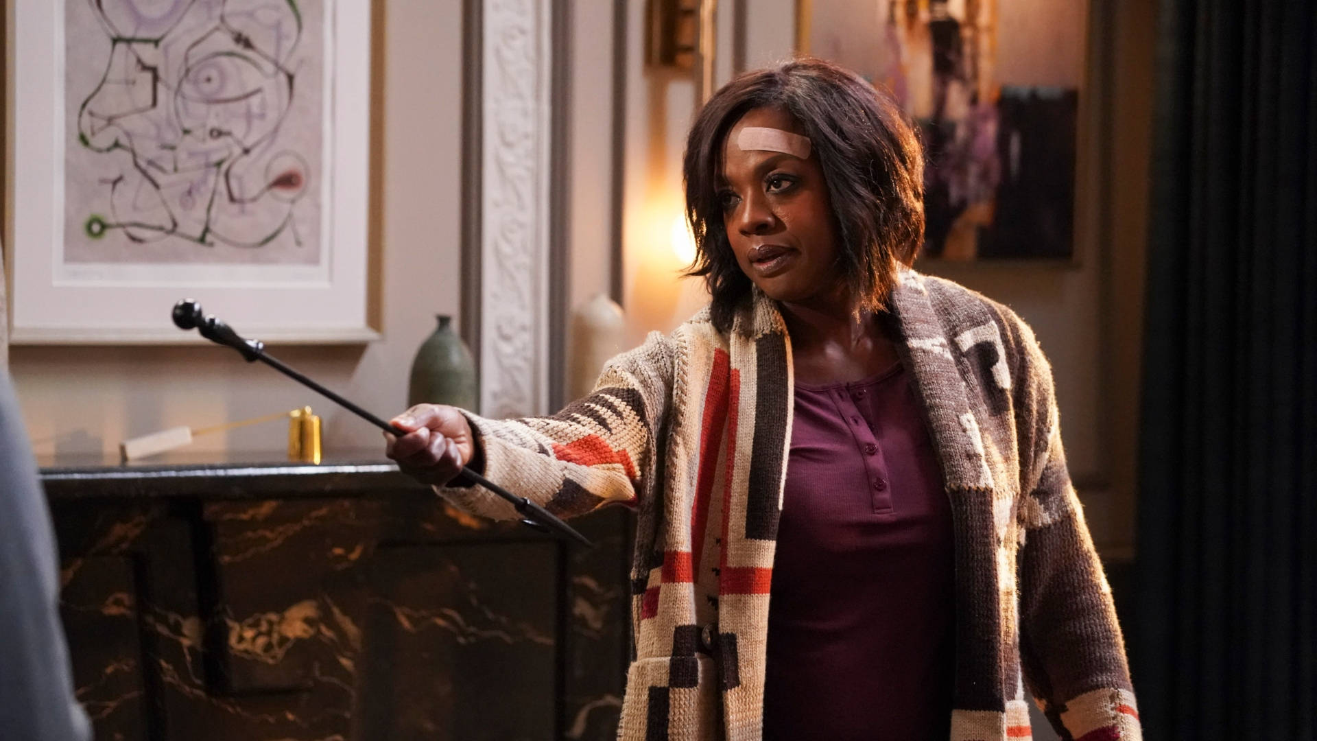 How To Get Away With Murder 桌布 壁紙 — 一位額頭纏著繃帶的女性，在一個溫暖照明、裝飾華麗的房間裡，手持魔杖，房間內有抽象藝術和大理石裝飾。