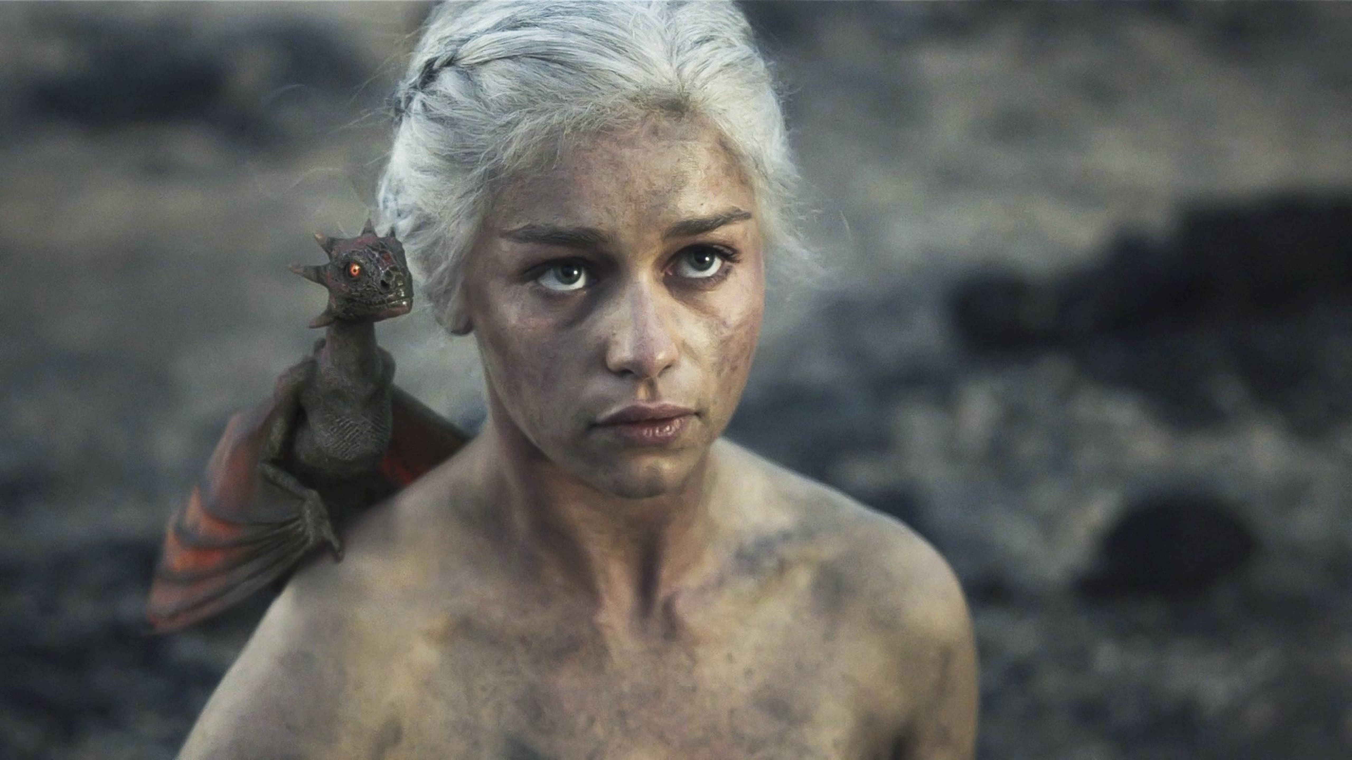 daenerys 桌布 壁紙 — 一位飽經戰鬥的白髮女子，肩上棲息著一條小龍，在岩石背景下凝視著。