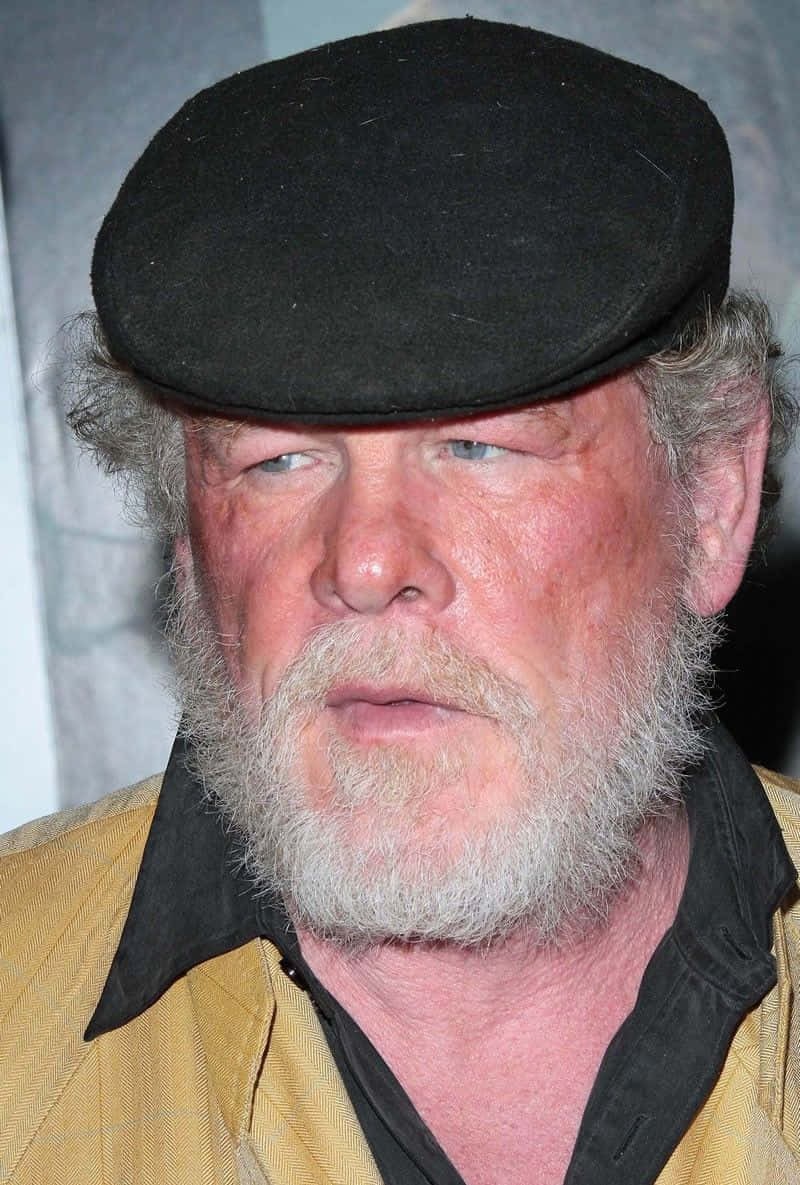 Nick Nolte 桌布 壁紙 — 一位飽經風霜、留著灰色鬍鬚、戴黑色平頂帽、穿黃色襯衫的男士特寫肖像。