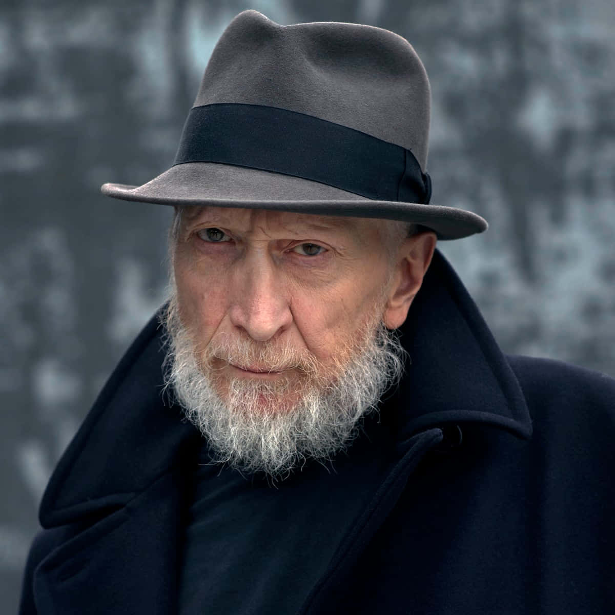 Frank Miller 桌布 壁紙 — 一位飽經風霜的白鬍子老人，戴著灰色軟呢帽和深色外套，背景是模糊的冬季森林。