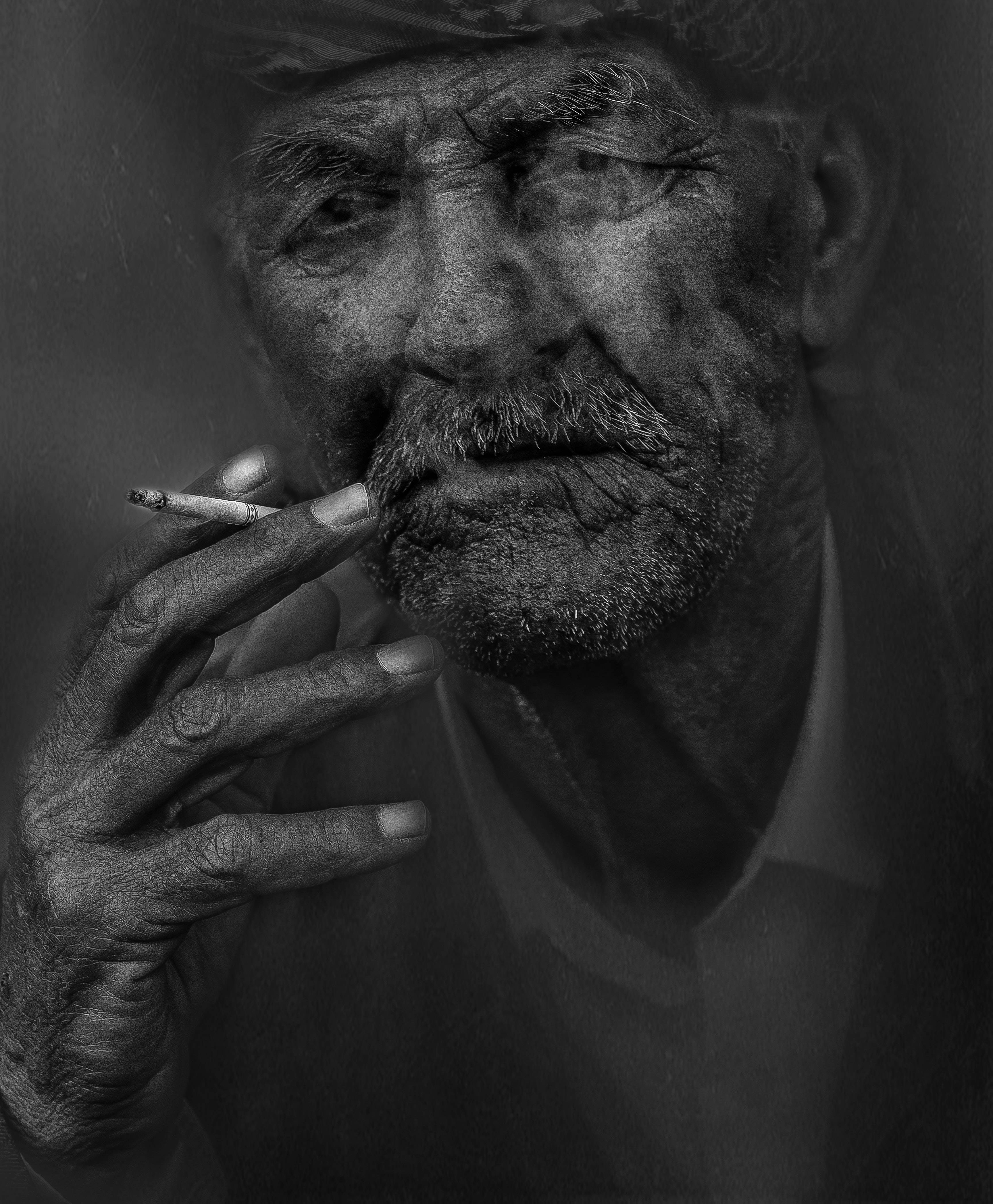 黑白 桌布 壁紙 — 一位飽經風霜的老人，臉上佈滿深深的皺紋，留著鬍鬚，在黑暗的背景中，用粗糙的手拿著一根香煙。