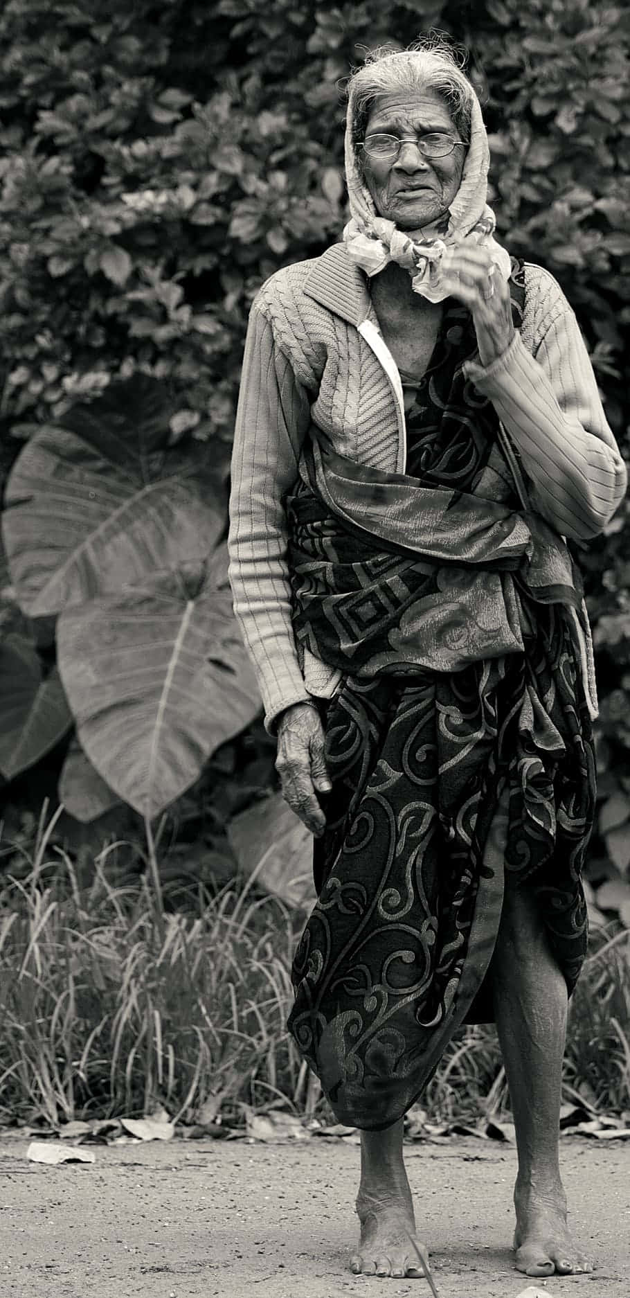 攝影 桌布 壁紙 — 一位飽經風霜的老年婦女穿著傳統服飾，赤腳站在一條被茂密樹葉環繞的泥路上，呈現黑白肖像。