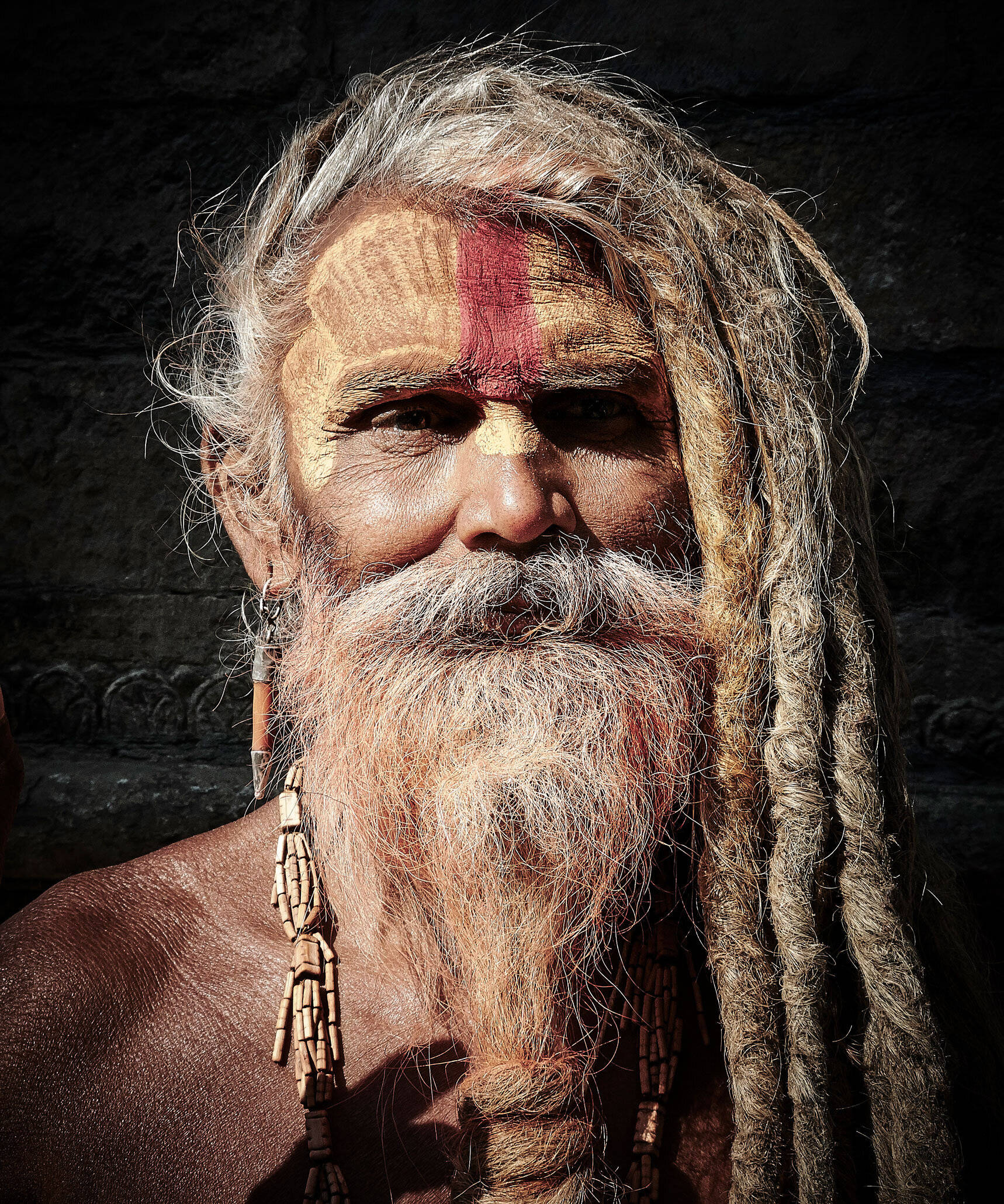 Aghori 桌布 壁紙 — 一位飽經風霜的聖人，留著長長的辮子，臉上塗著神聖的紅色和金色顏料，在黑暗的石牆前凝視著。