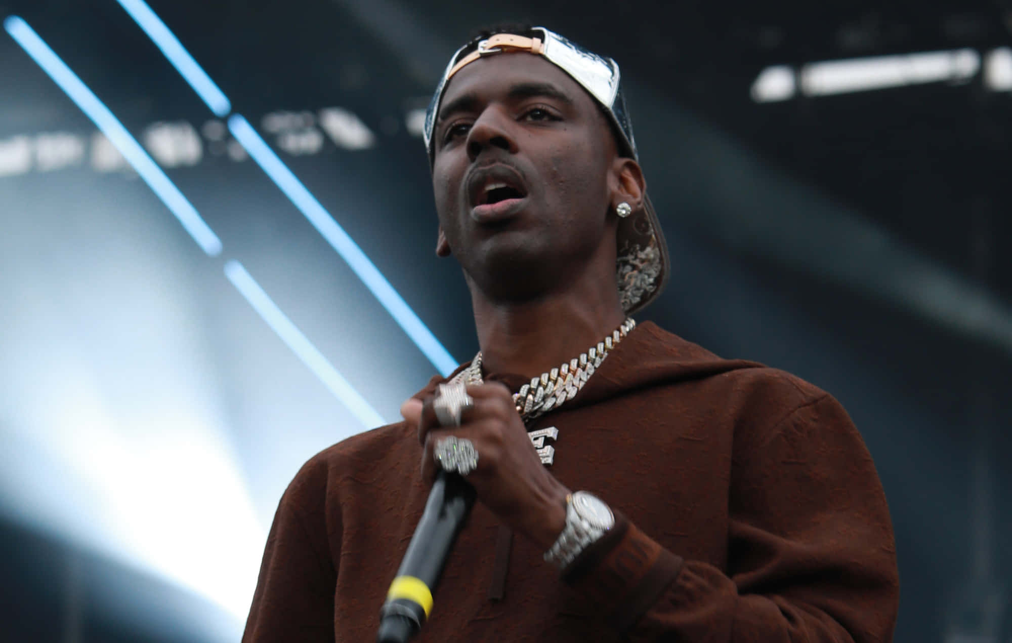 Young Dolph 桌布 壁紙 — 一位饒舌歌手在明亮的藍色舞台燈光下，穿著棕色連帽衫和金色鏈條，熱情地表演。