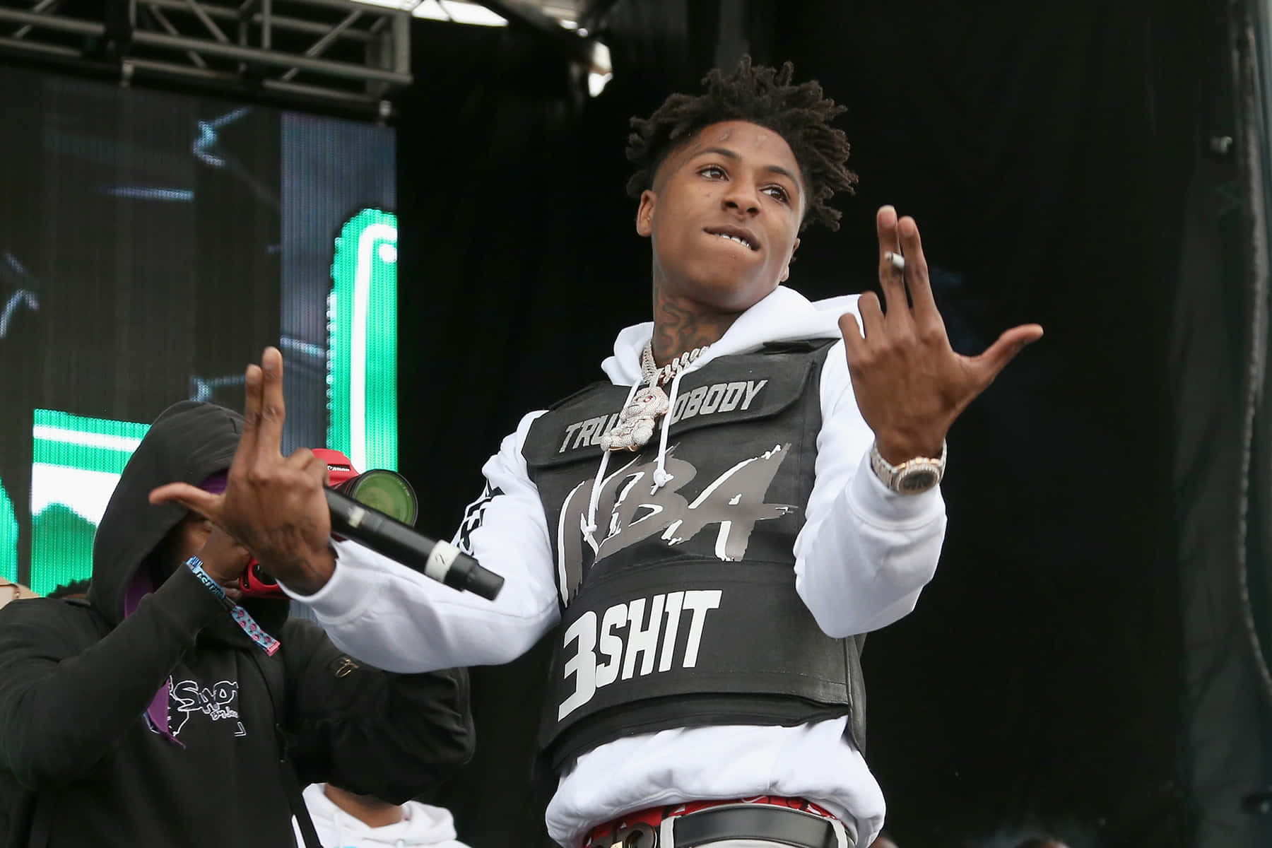 Youngboy 桌布 壁紙 — 一位饒舌歌手在舞台上充滿活力地表演，身穿白色連帽衫，外搭一件帶有粗體白色文字的黑色背心。