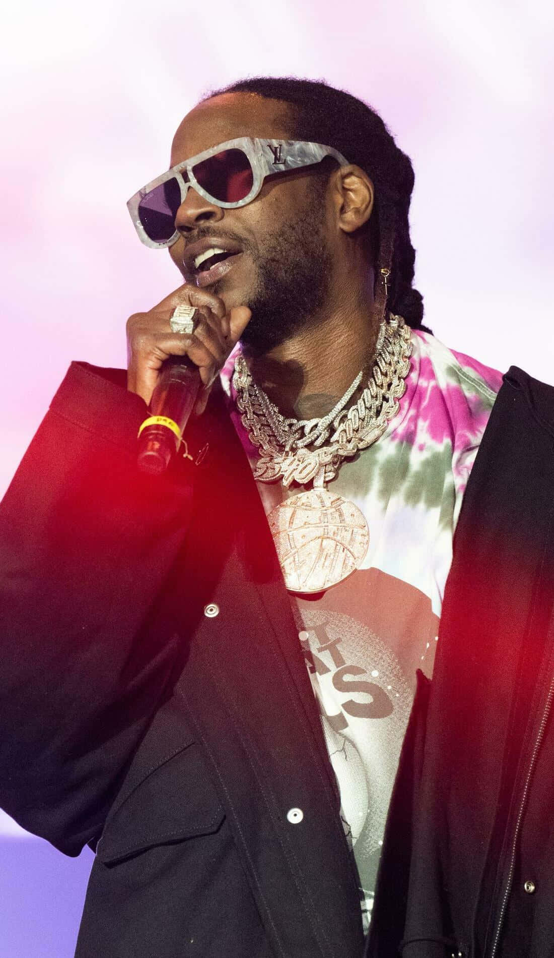 2 Chainz 桌布 壁紙 — 一位饒舌歌手戴著超大太陽眼鏡和層疊鏈條，在充滿活力的舞台燈光下對著麥克風表演。