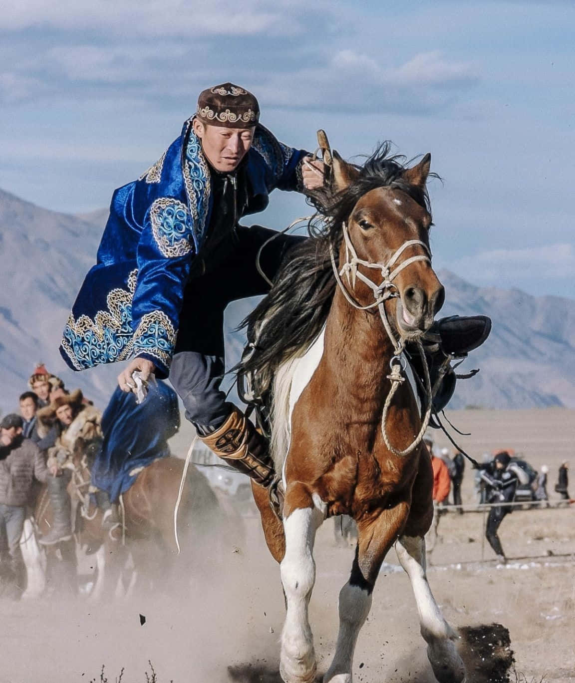 Mongolian 桌布 壁紙 — 一位騎手身穿華麗的藍色傳統服飾，在一匹奔騰的棕白相間的馬上俯身前傾，馬蹄揚起塵土，背景是山巒疊嶂。