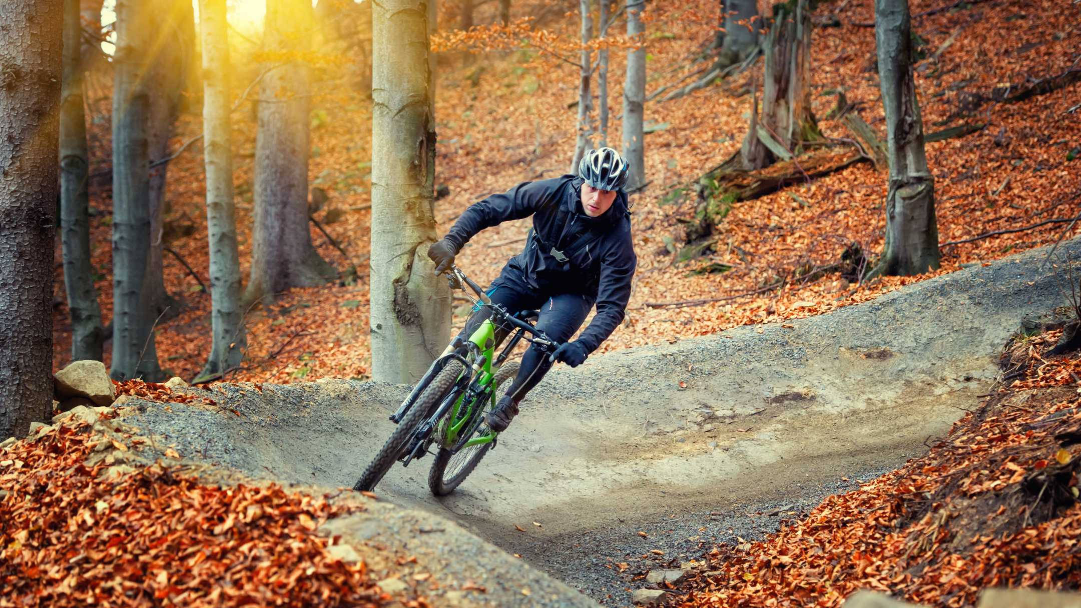 Mountain Bike 桌布 壁紙 — 一位騎自行車的人在鋪滿金色秋葉的森林小徑上傾身過彎，陽光穿過樹林灑落。
