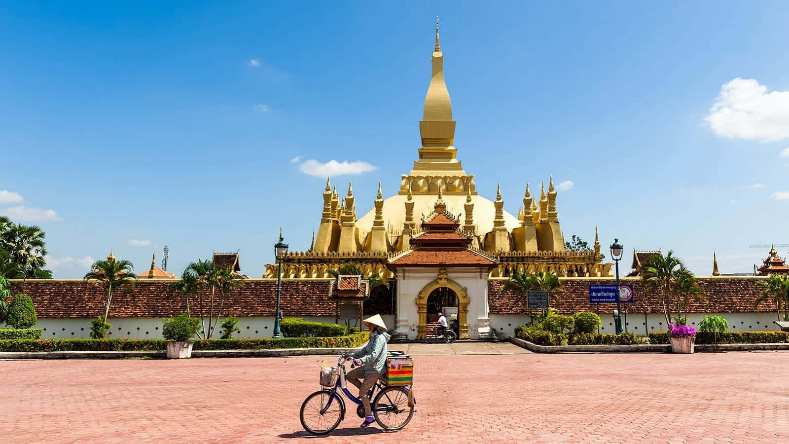 Laos 桌布 壁紙 — 一位騎自行車的人踩過一座宏偉的金色寺廟，天空湛藍，白雲點綴。