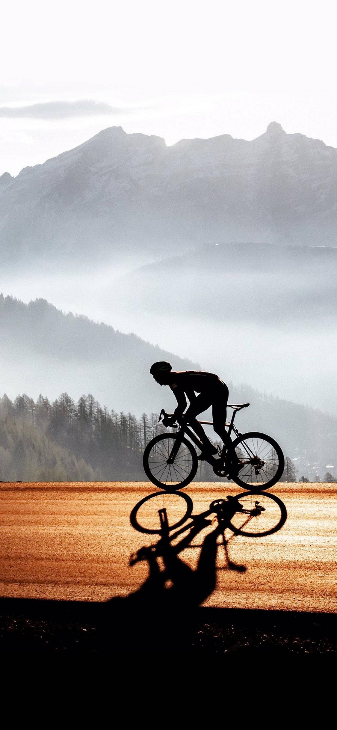 Mountain Bike 桌布 壁紙 — 一位騎自行車者的剪影穿過霧氣瀰漫的山谷，在潮濕的地面上投下長長的戲劇性影子。
