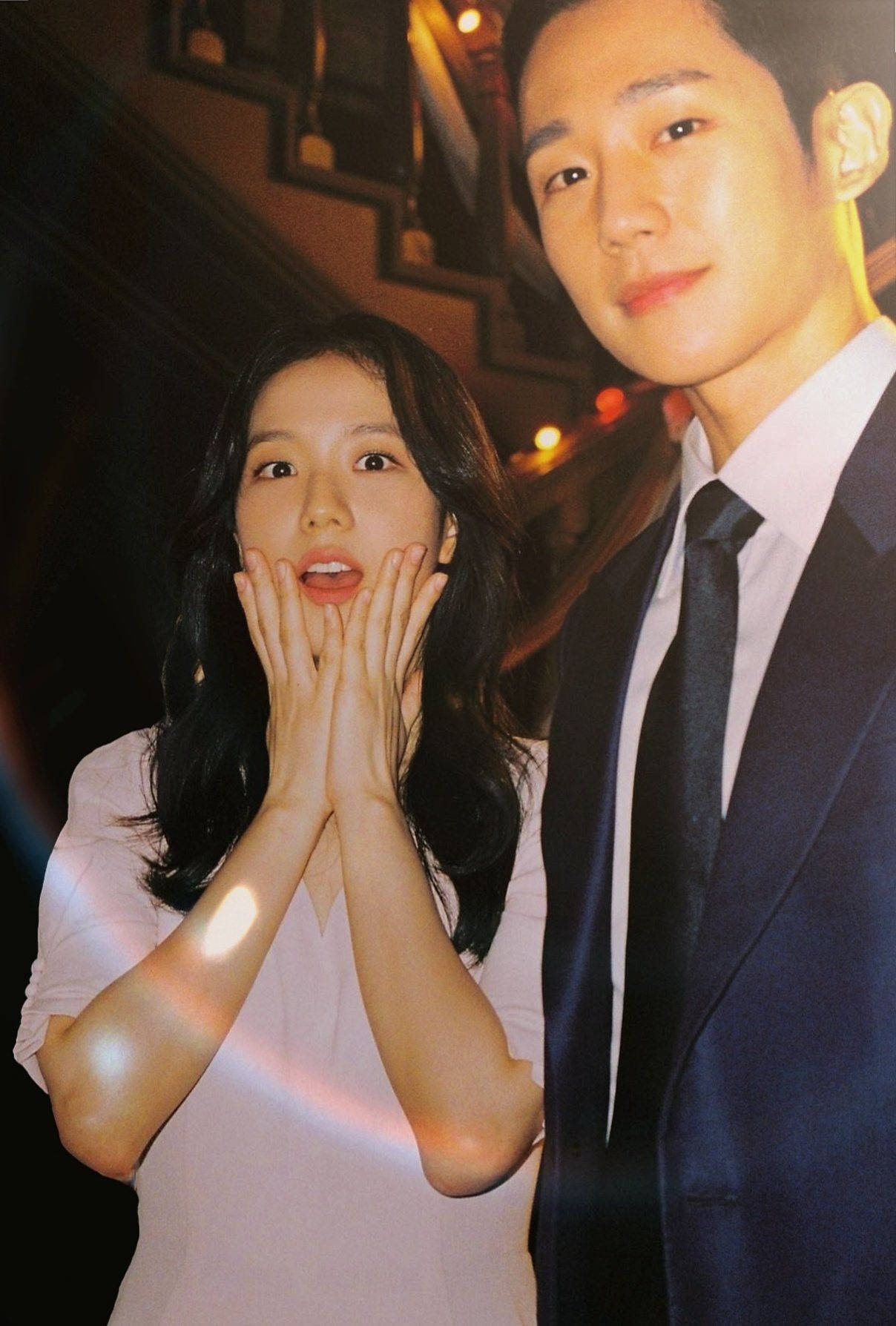 drama 桌布 壁紙 — 一位驚訝的女士雙手放在臉頰上，站在一位穿著西裝微笑的男士旁邊，在溫暖的環境燈光下。