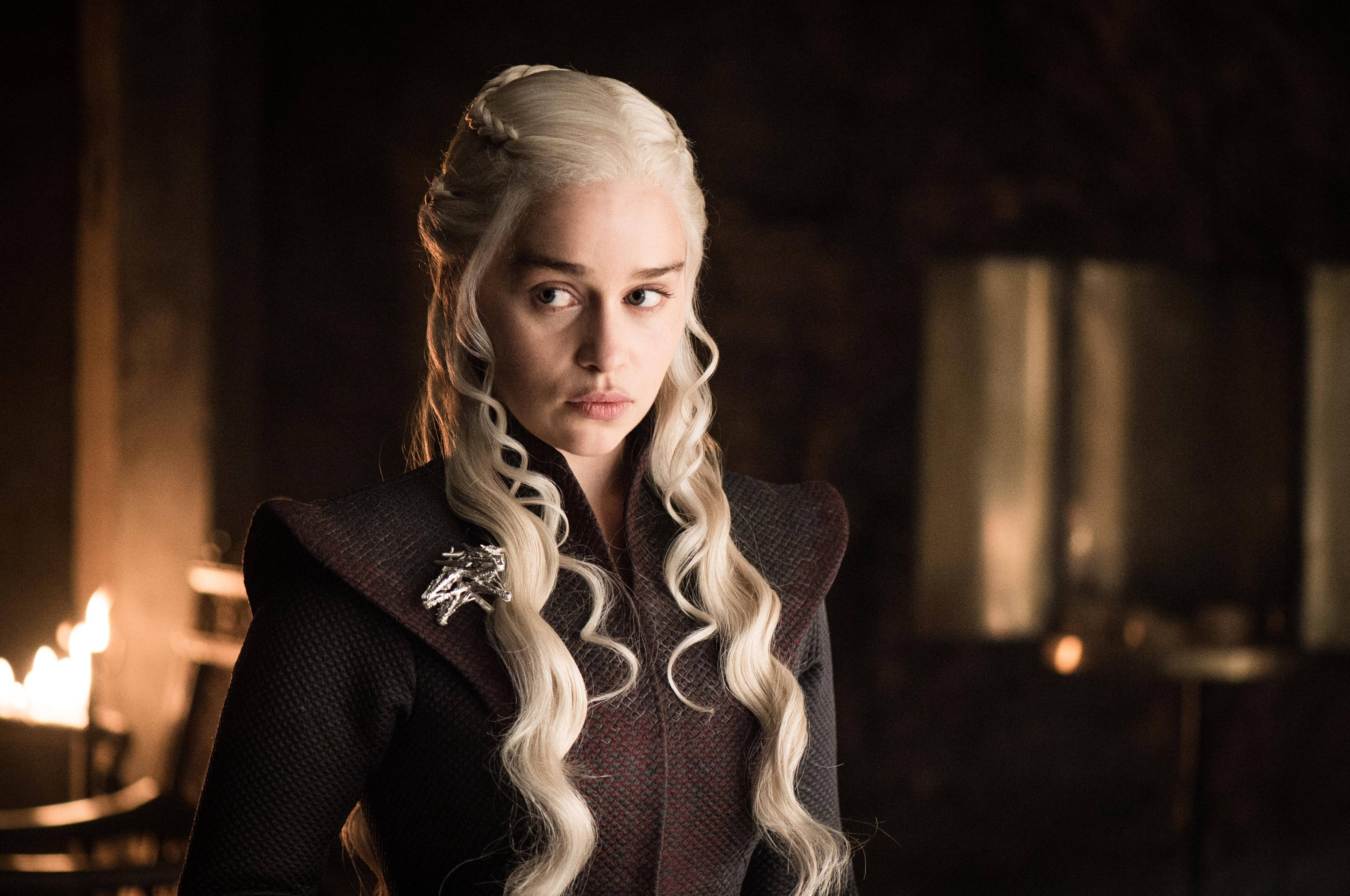 daenerys 桌布 壁紙 — 一位高貴的女性，留著長長的鉑金色頭髮，戴著龍形胸針，在燭光搖曳的昏暗房間裡專注地凝視。