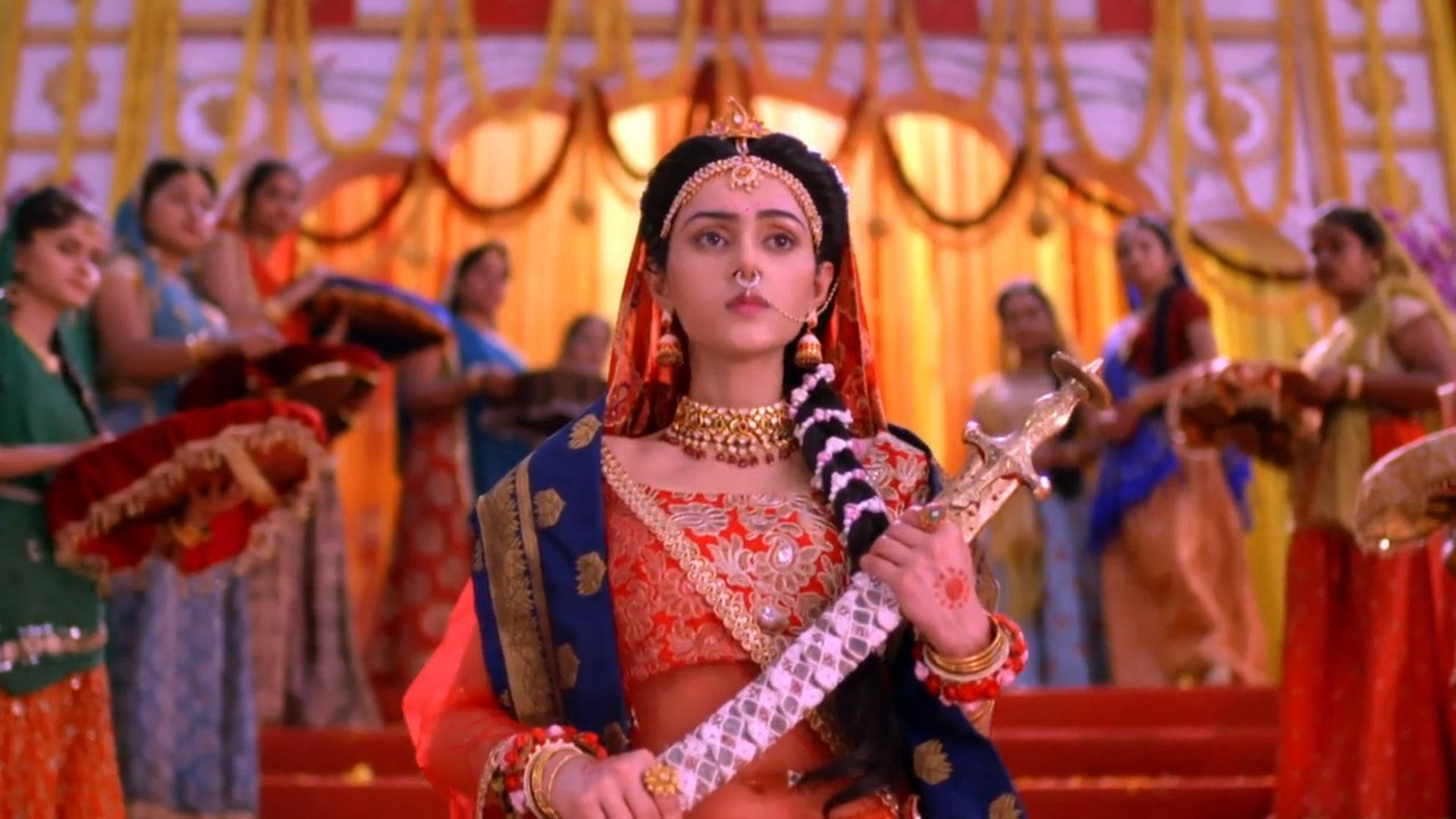 radha krishna serial 桌布 壁紙 — 一位高貴的女性穿著華麗的傳統印度服飾，在充滿活力的儀式場景中，被舞者包圍，手持一把劍。