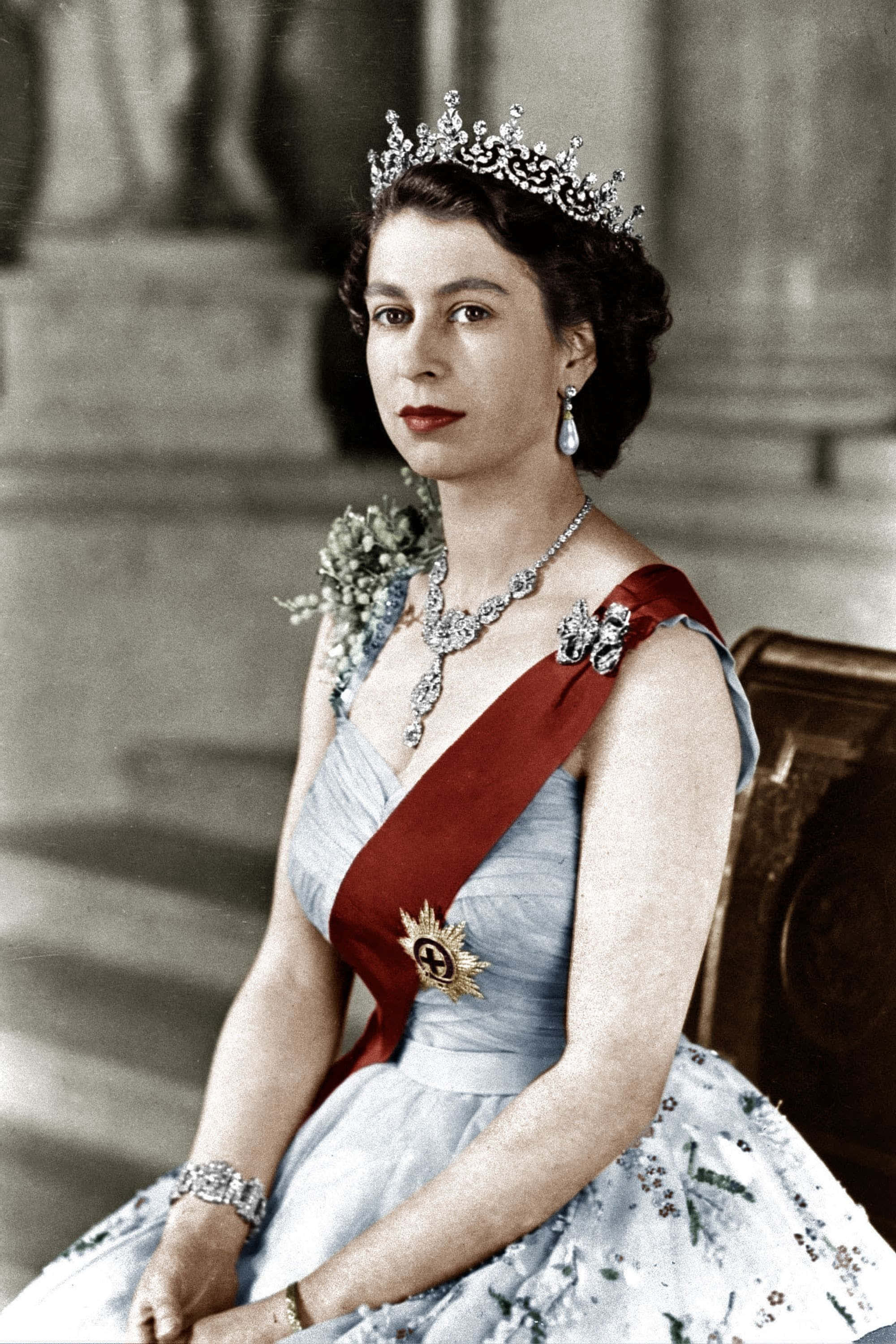 Queen Elizabeth 桌布 壁紙 — 一位高貴的女性身穿淺藍色長袍，飾有鑽石和紅色腰帶，戴著王冠和珍珠耳環，端莊地坐著。