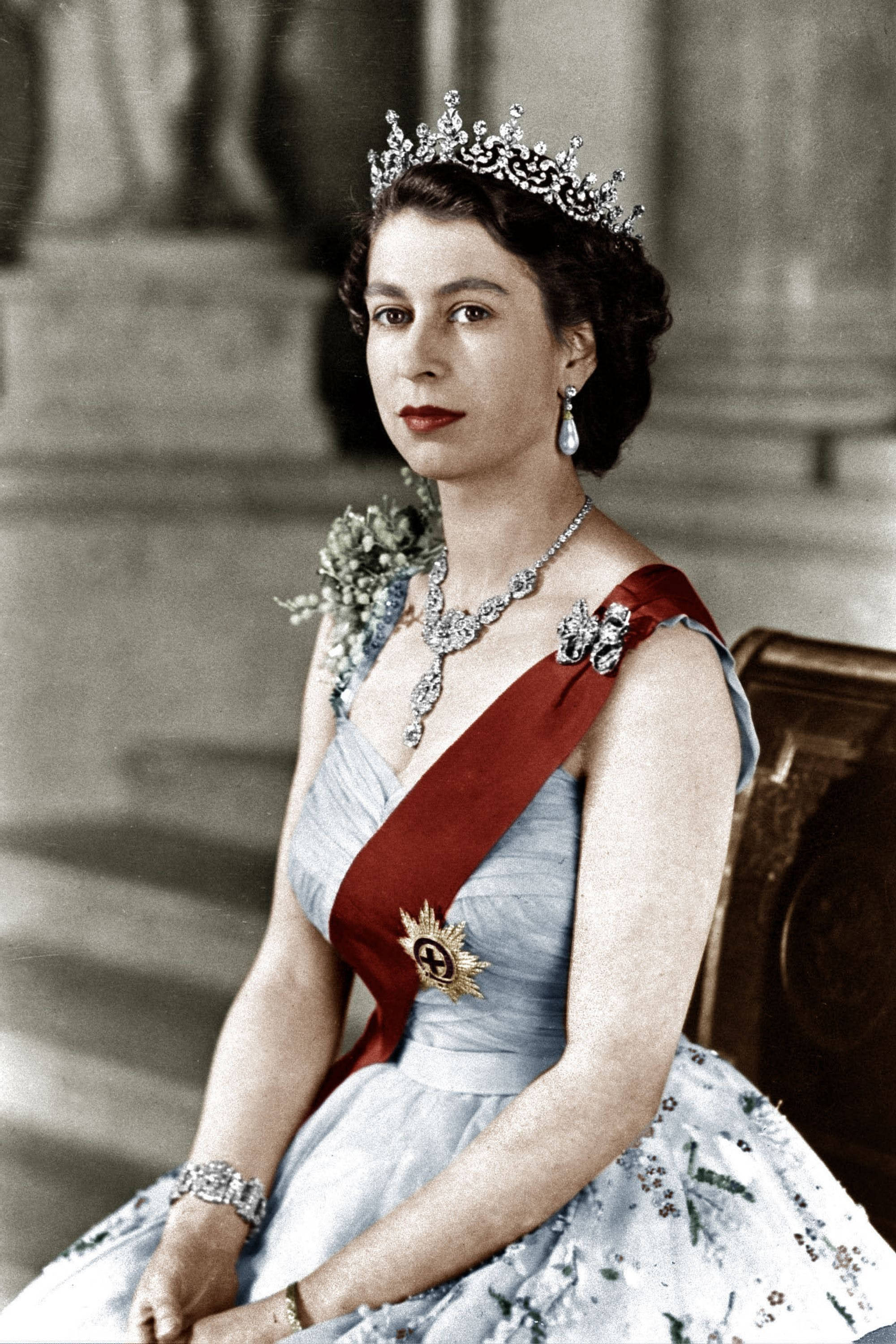 Queen Elizabeth 桌布 壁紙 — 一位高貴的女性身穿淺藍色長袍，飾有珠寶和紅色腰帶，端莊地坐著，戴著閃閃發光的頭飾和珍珠耳環。