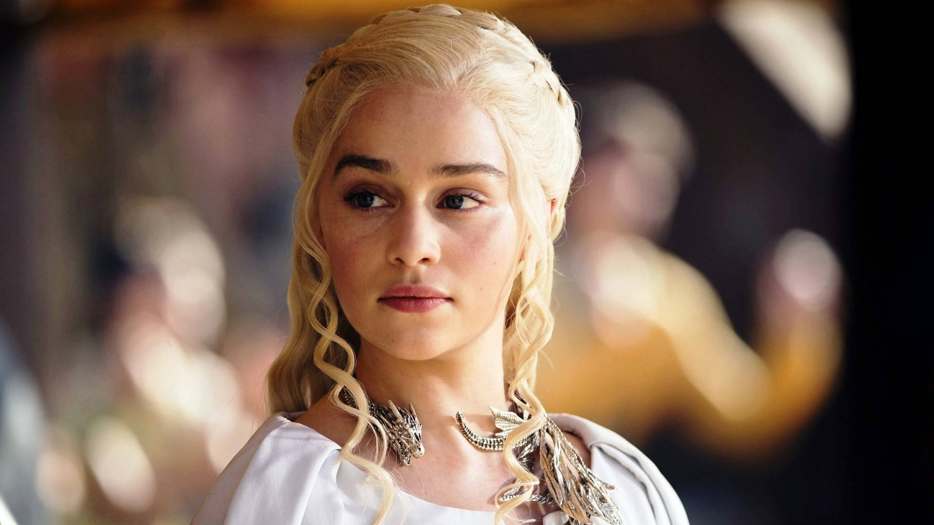 Emilia 桌布 壁紙 — 一位高貴的金髮女子，編著辮子，佩戴龍主題珠寶，在溫暖的室內環境中專注地凝視。