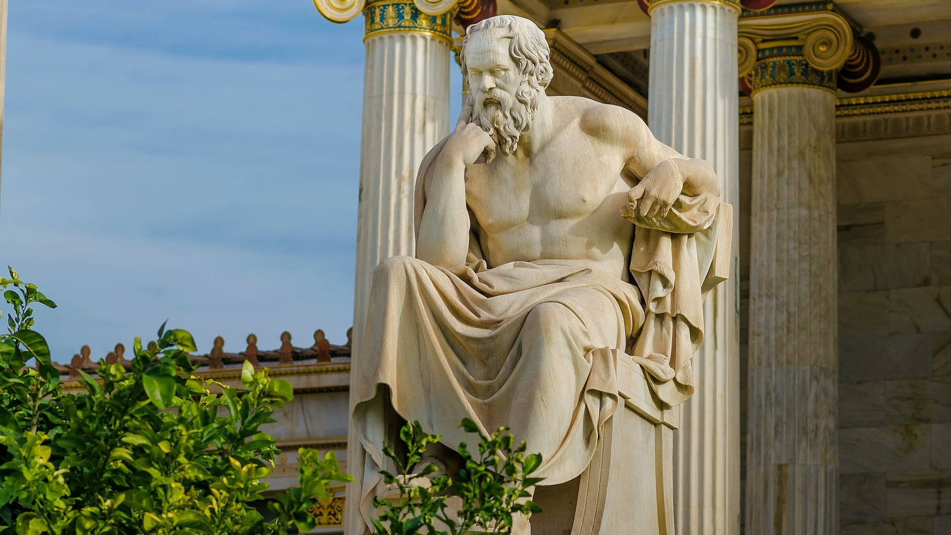 Greek Aesthetic 桌布 壁紙 — 一位鬍鬚哲學家的大理石雕像，坐在裝飾華麗的柱子下沉思，前景有綠色樹葉。
