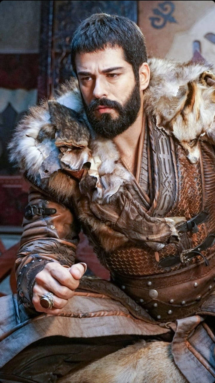 Ertugrul 桌布 壁紙 — 一位鬍鬚戰士身穿毛皮襯裡盔甲，手持匕首，兩隻狼棲息在他的肩膀上，處於燈光昏暗的中世紀大廳中。