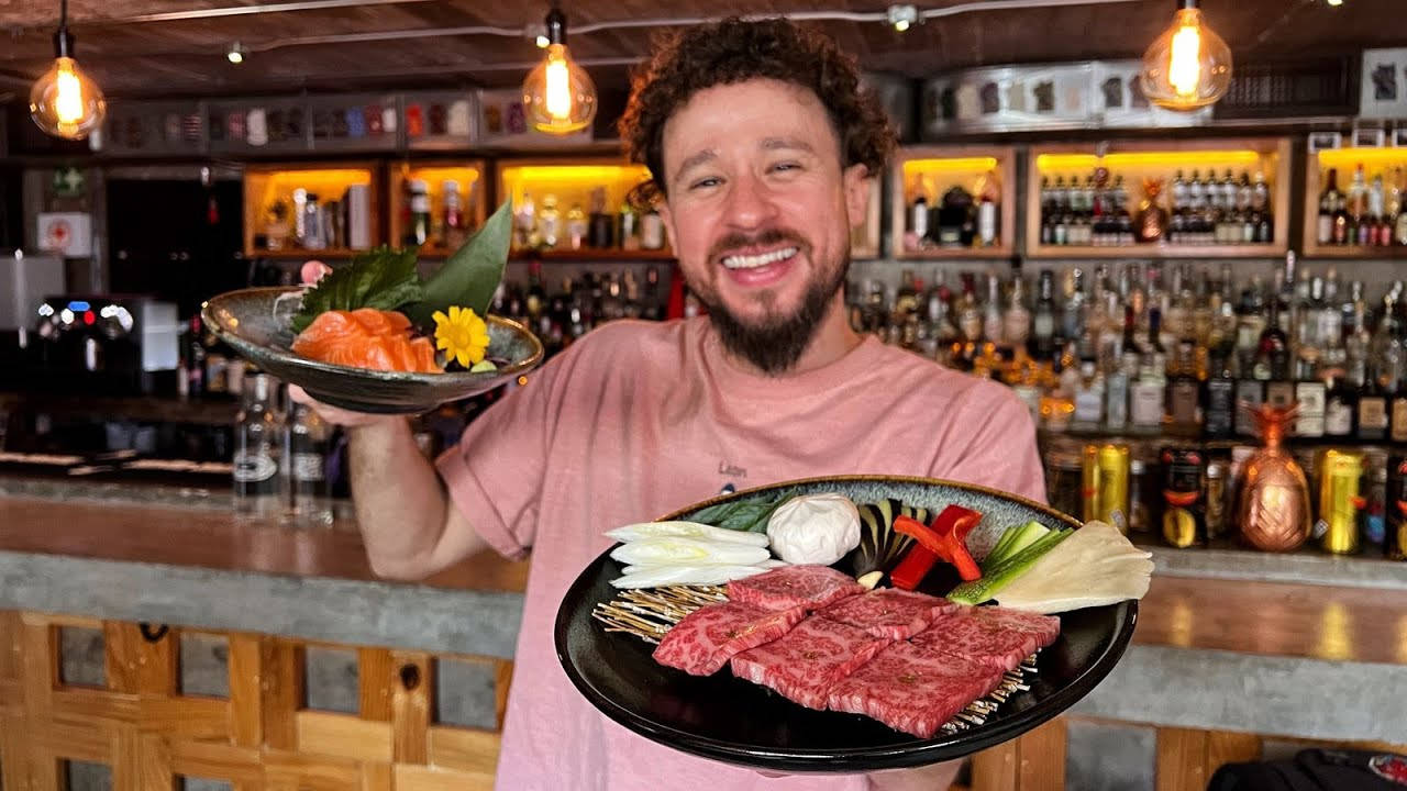 Restaurant 桌布 壁紙 — 一位鬍鬚男在溫暖燈光的酒吧裡，自豪地展示兩盤刺身和烤肉，笑容滿面。