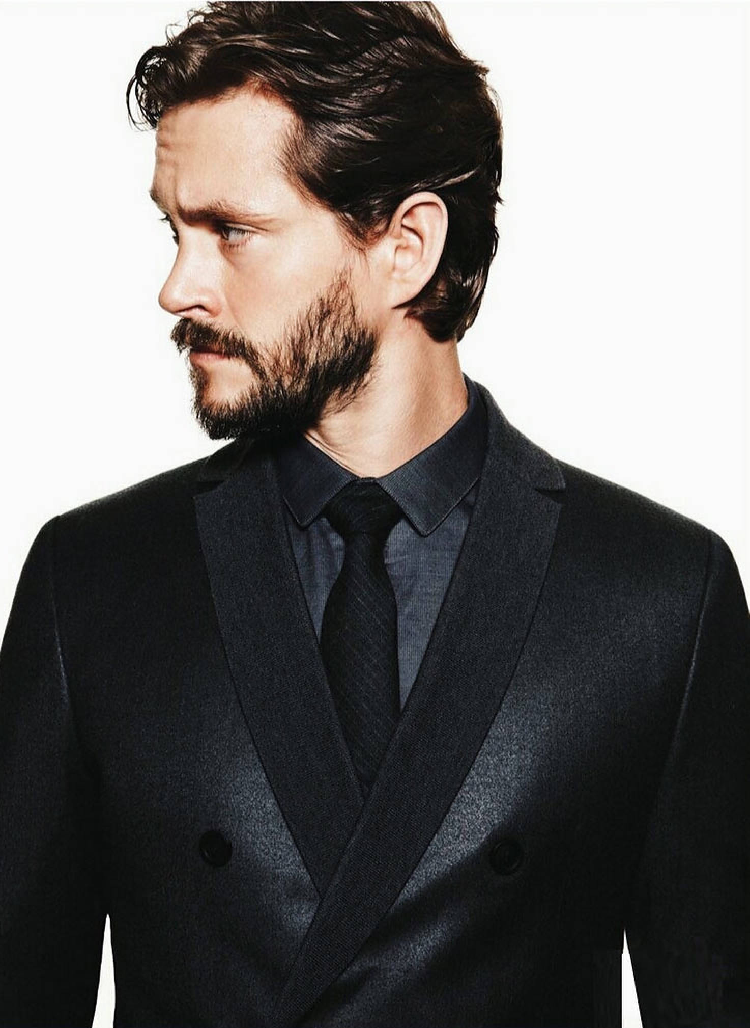 Hugh Dancy 桌布 壁紙 — 一位鬍鬚男子身穿深色西裝，在純白背景下若有所思地凝視。