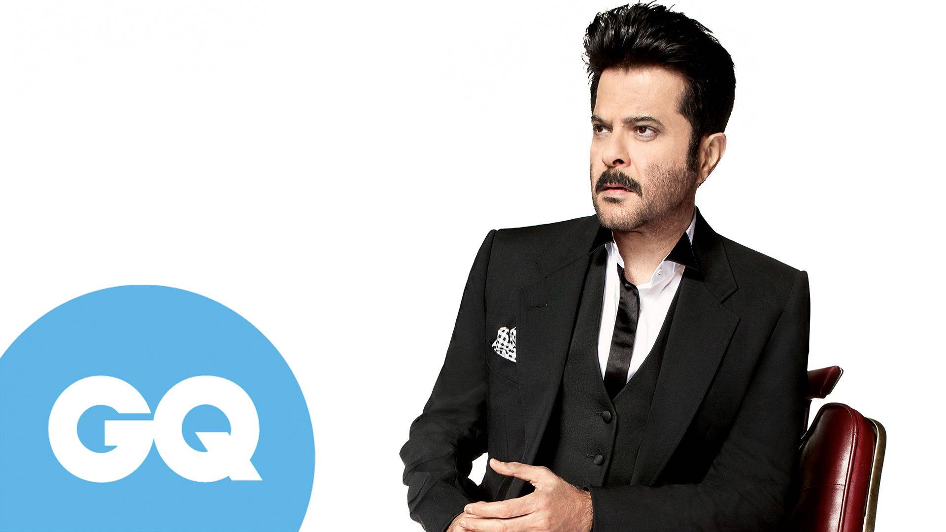 anil kapoor 桌布 壁紙 — 一位鬍鬚男穿著時尚，自信地坐在白色背景的紅色椅子上，帶有GQ標誌。
