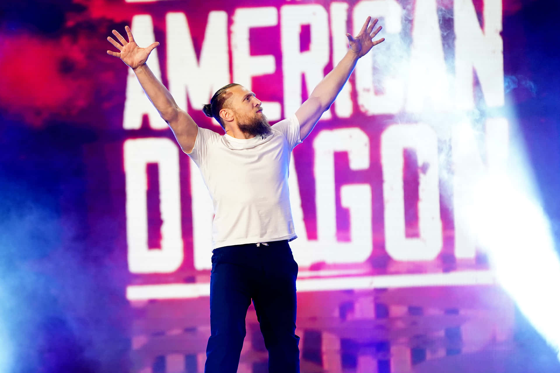 摔角 桌布 壁紙 — 一位鬍鬚男穿著白色T恤，在明亮的燈光和「AMERICAN DRAGON」背景下手舉雙臂，勝利地站在舞台上。