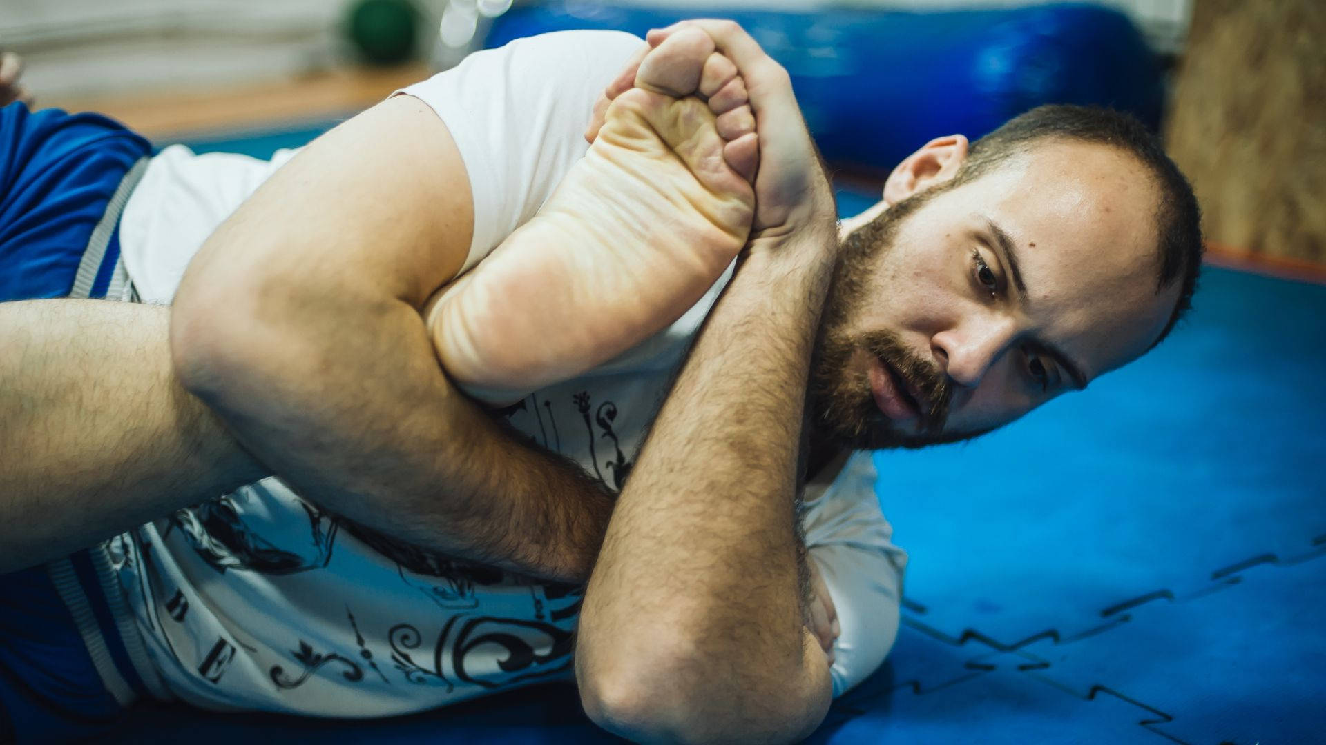 martial arts 桌布 壁紙 — 一位鬍鬚男穿著白襯衫，在藍色健身墊上專注地進行深層坐姿伸展。