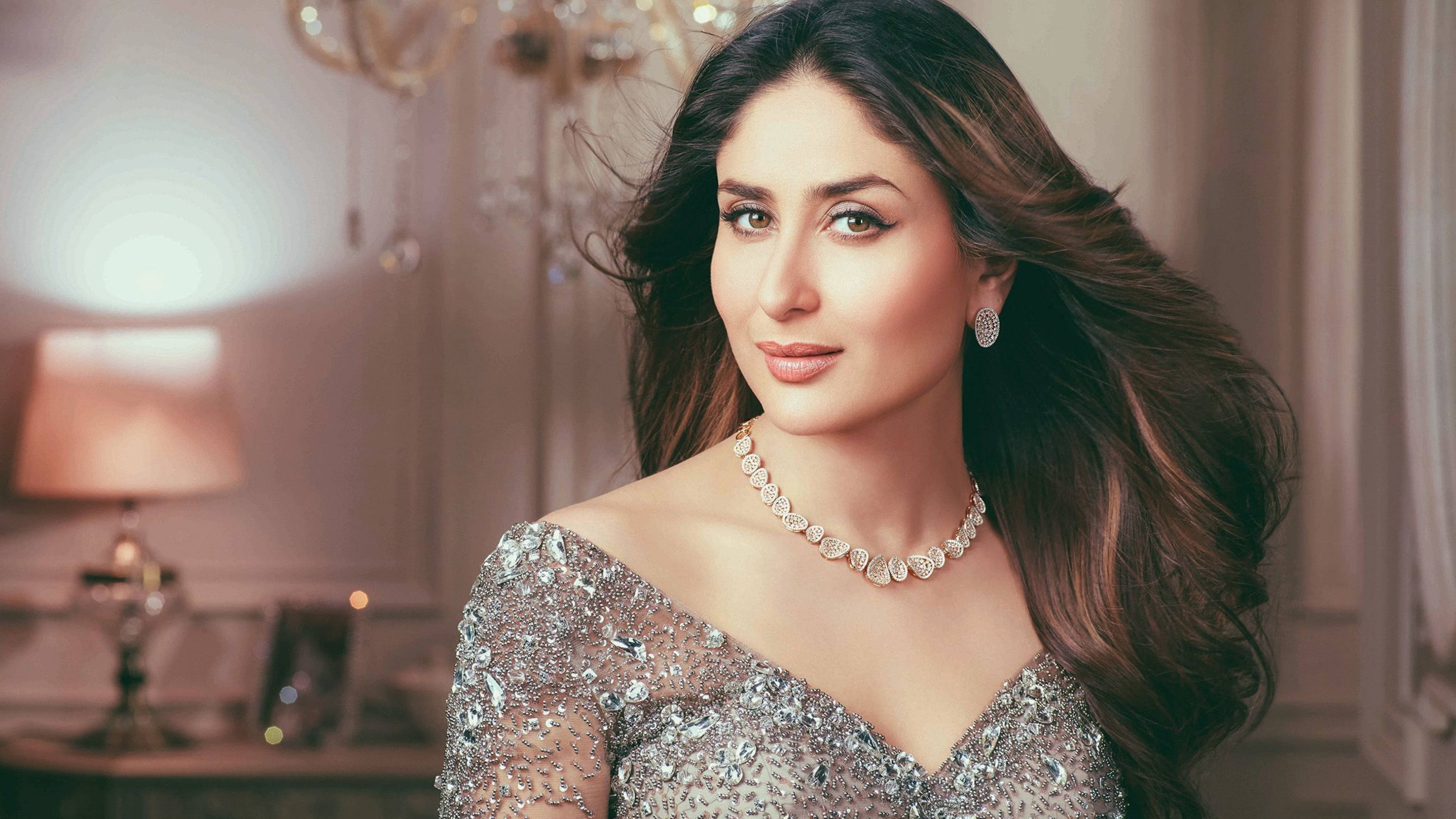 Kareena 桌布 壁紙 — 一位魅力四射、擁有飄逸深色長髮的女性，在柔和燈光的優雅房間中，身穿閃耀的銀色禮服並佩戴鑽石珠寶。