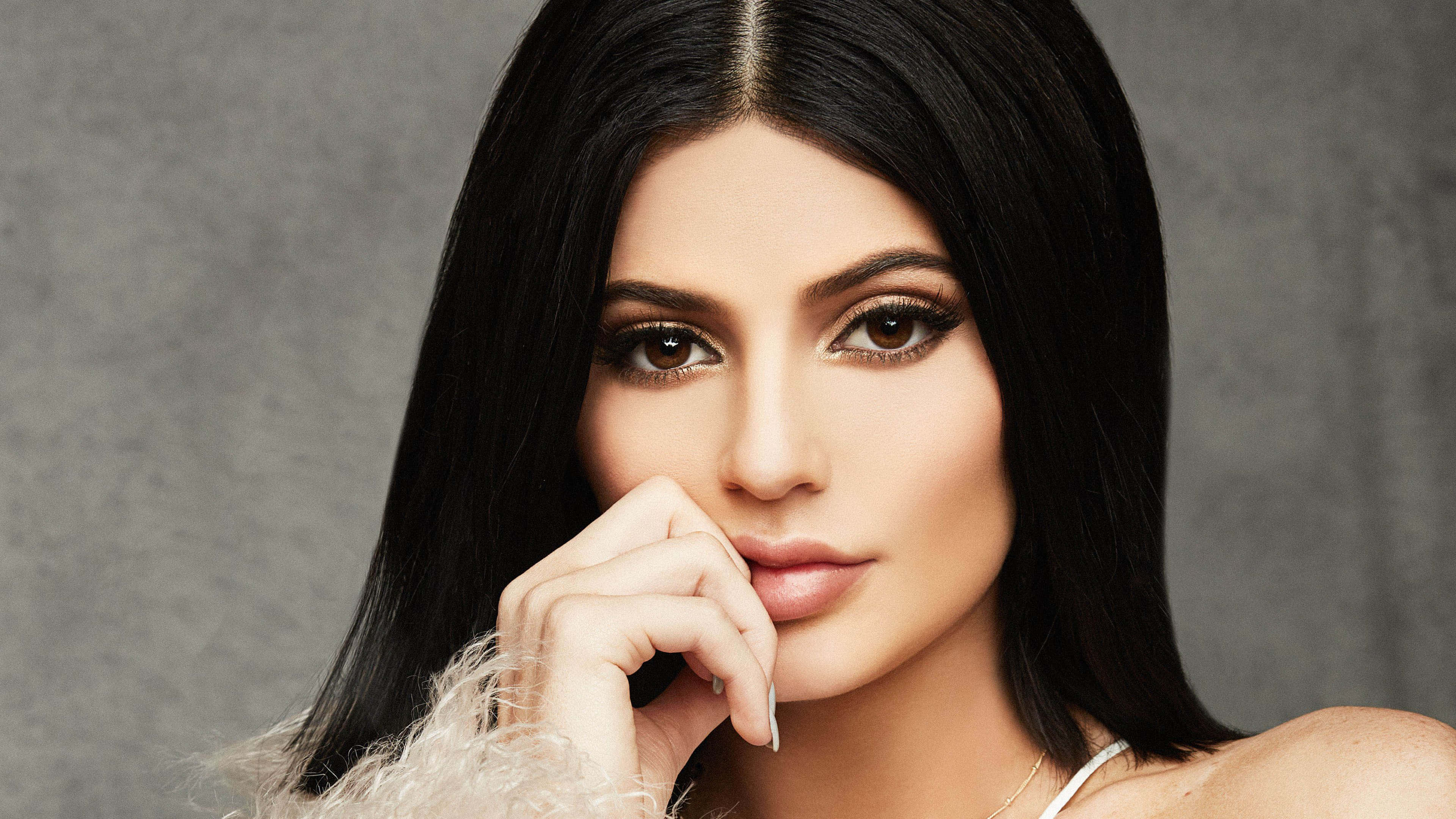 Kylie Jenner 4K 桌布 壁紙 — 一位魅力四射的女士，長長的黑髮和煙燻眼妝，在灰色紋理背景前將手靠近嘴唇擺姿勢。