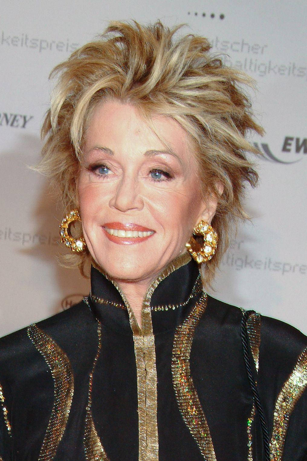 Celebrity 桌布 壁紙 — 一位魅力四射的女性，擁有豐盈的金色頭髮和金色圓環耳環，穿著黑色和金色裝飾的服裝，溫暖地微笑。