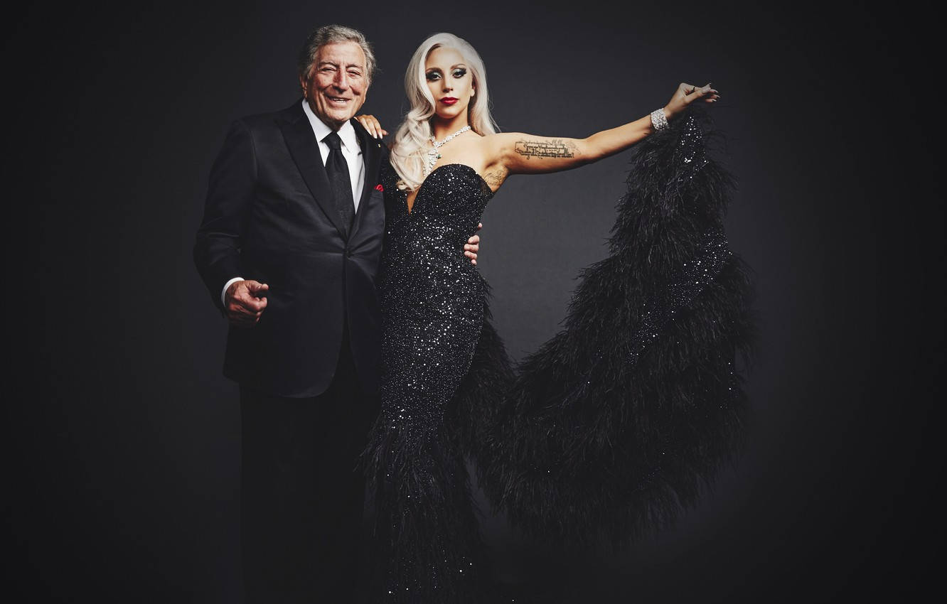 Lady Gaga 桌布 壁紙 — 一位魅力四射的男士身穿西裝，一位耀眼奪目的女士身穿黑色羽毛禮服，在深色工作室背景前自信地擺姿勢。