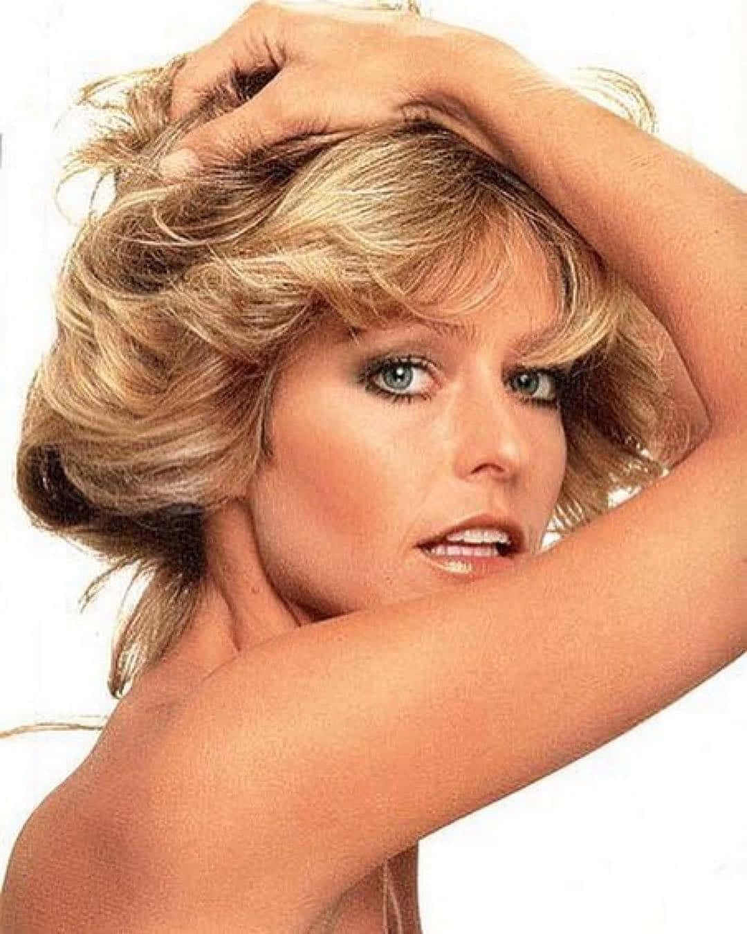 Farrah Fawcett 桌布 壁紙 — 一位魅力四射的金髮女郎，擁有豐盈的秀髮和引人注目的藍色眼睛，在純白背景前自信地擺姿勢。