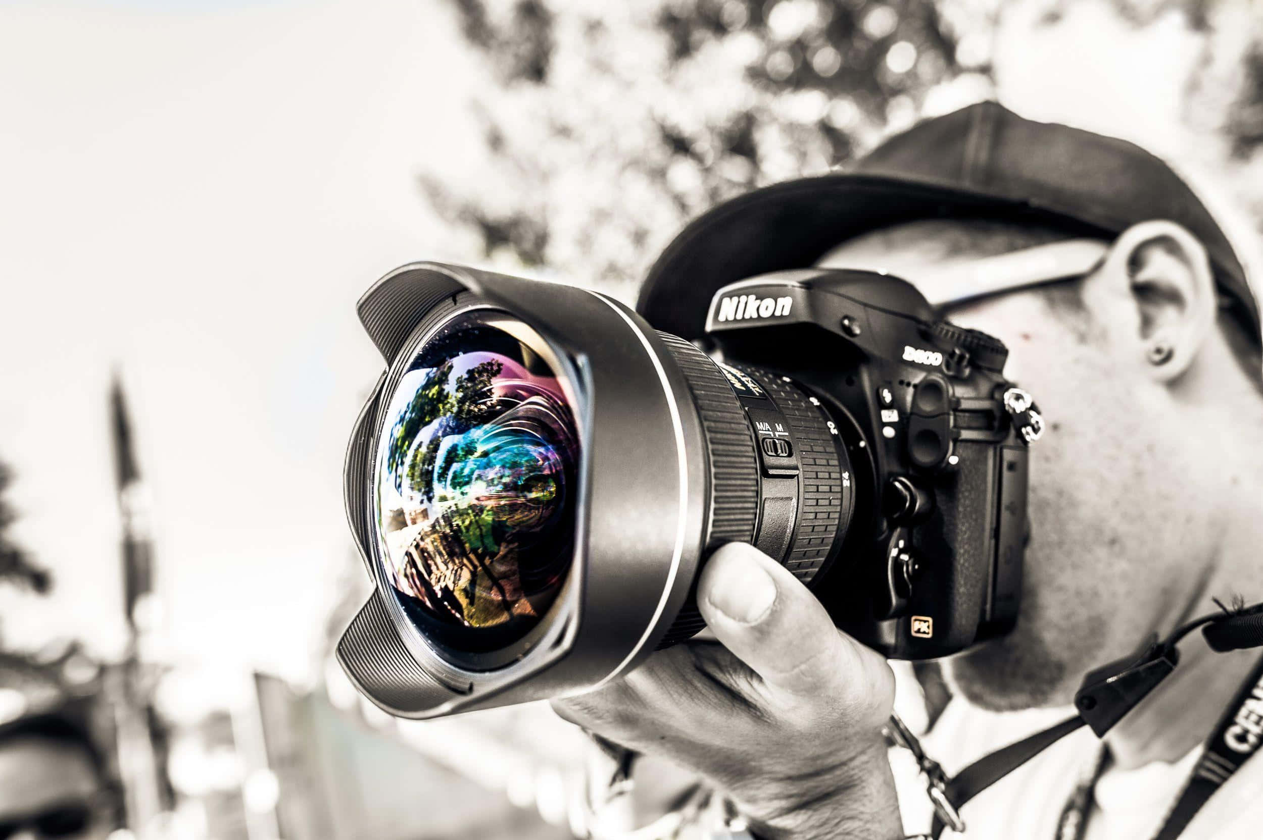 攝影師 桌布 壁紙 — 一位黑白攝影師手持一部Nikon DSLR相機，鏡頭中有色彩繽紛的彩虹倒影。