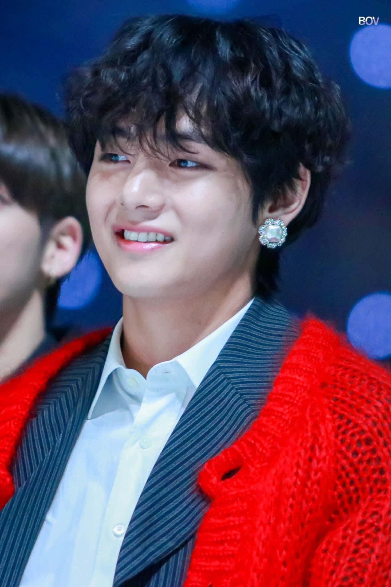 Taehyung Cute 桌布 壁紙 — 一位黑色捲髮的年輕男子，穿著紅色針織開襟衫搭配條紋西裝和閃亮耳環，露出溫暖的笑容。