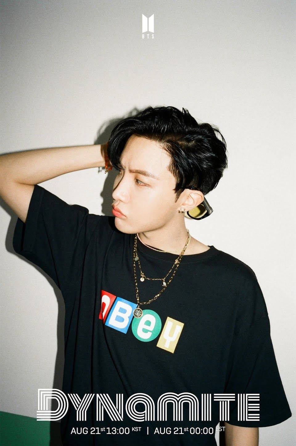jhope 桌布 壁紙 — 一位黑髮、佩戴大膽珠寶的年輕男子，穿著印有彩色字母的黑色T恤，自信地靠在素牆上擺姿勢。