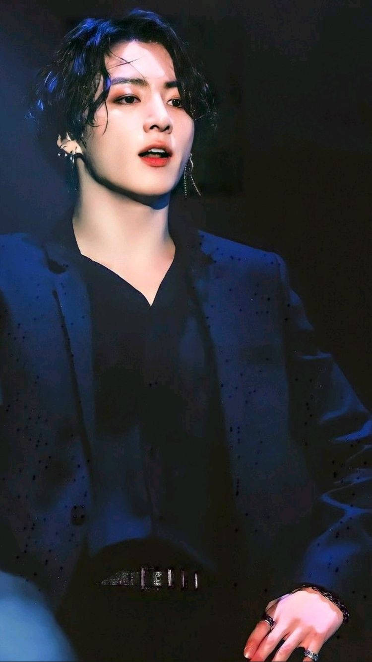 Jungkook 桌布 壁紙 — 一位黑髮、妝容出眾的年輕男子，在戲劇性的舞台燈光下穿著藍色西裝外套。
