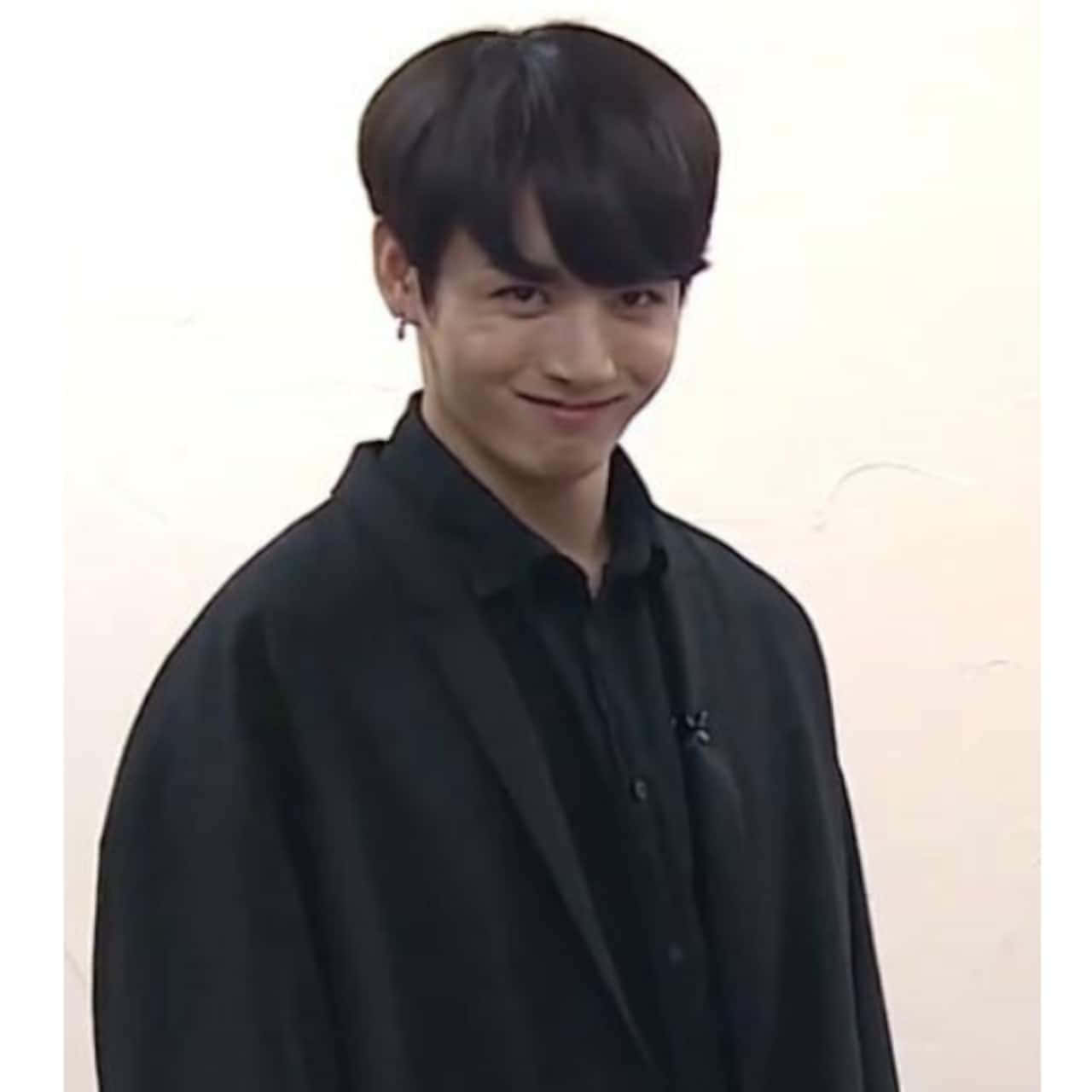 BTS 桌布 壁紙 — 一位黑髮、帶著微妙微笑的年輕男子，穿著黑色鈕扣襯衫，背景是純色的米白色牆壁。