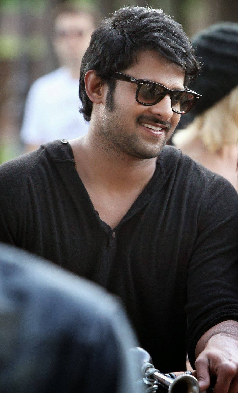 Prabhas Mirchi 桌布 壁紙 — 一位黑髮、戴太陽眼鏡的男士微笑著騎摩托車穿過模糊的戶外場景。