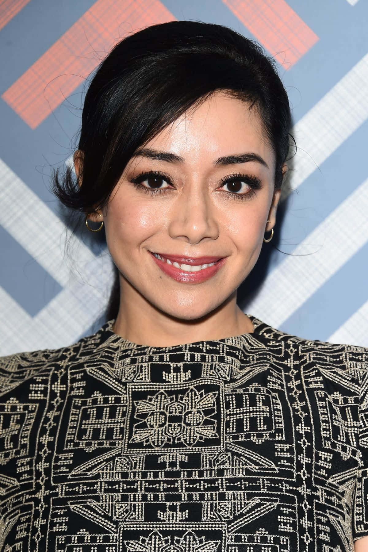 Aimee Garcia 桌布 壁紙 — 一位黑髮、戴金色耳環的微笑女性，身穿黑金圖案上衣，背景為藍橙幾何圖案。