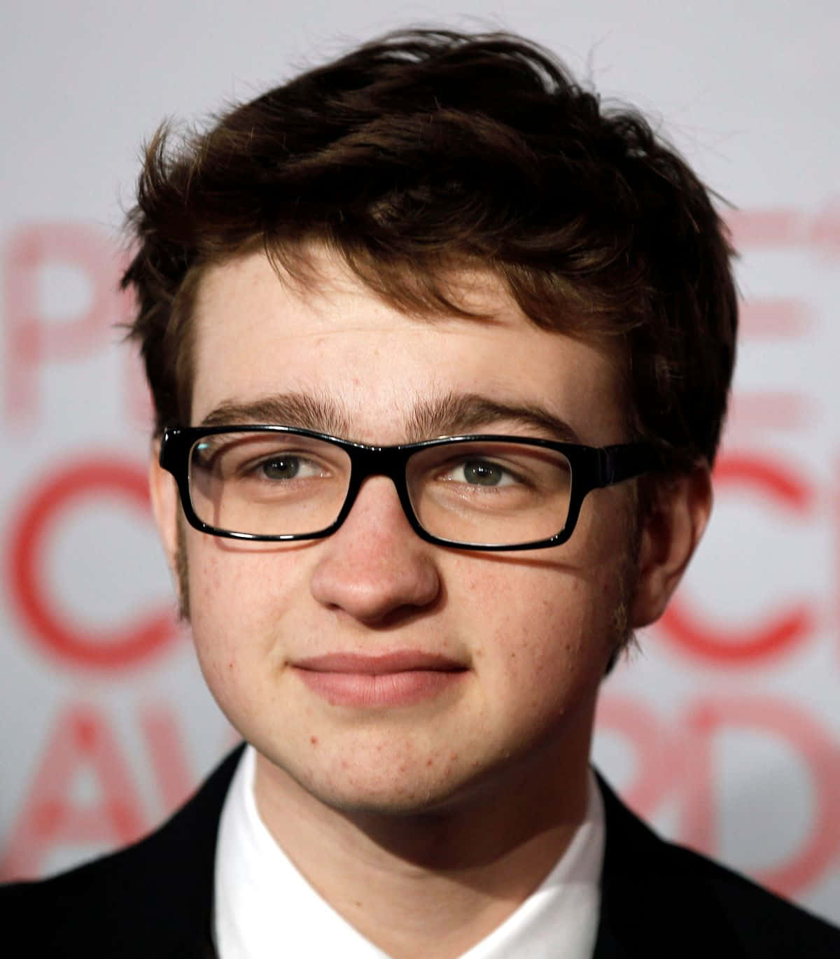 actor 桌布 壁紙 — 一位黑髮、戴黑色框眼鏡的年輕男子，穿著黑色西裝和白色襯衫，在模糊的紅白背景下溫柔微笑。