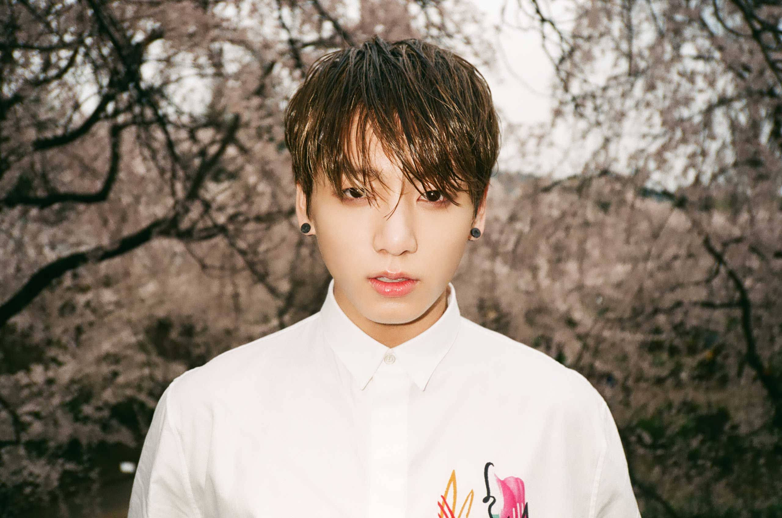 Jungkook 桌布 壁紙 — 一位黑髮、戴黑色耳擴的年輕男子，穿著帶有彩色刺繡的白襯衫，以盛開的櫻花樹為背景，直視鏡頭。