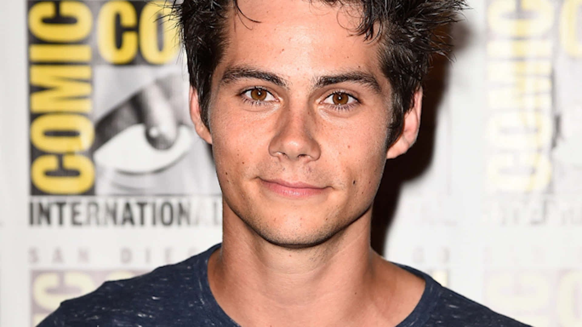 Dylan Obrien 桌布 壁紙 — 一位黑髮、溫暖棕色眼睛的年輕男子對著鏡頭溫柔微笑，背景是Comic-Con International。