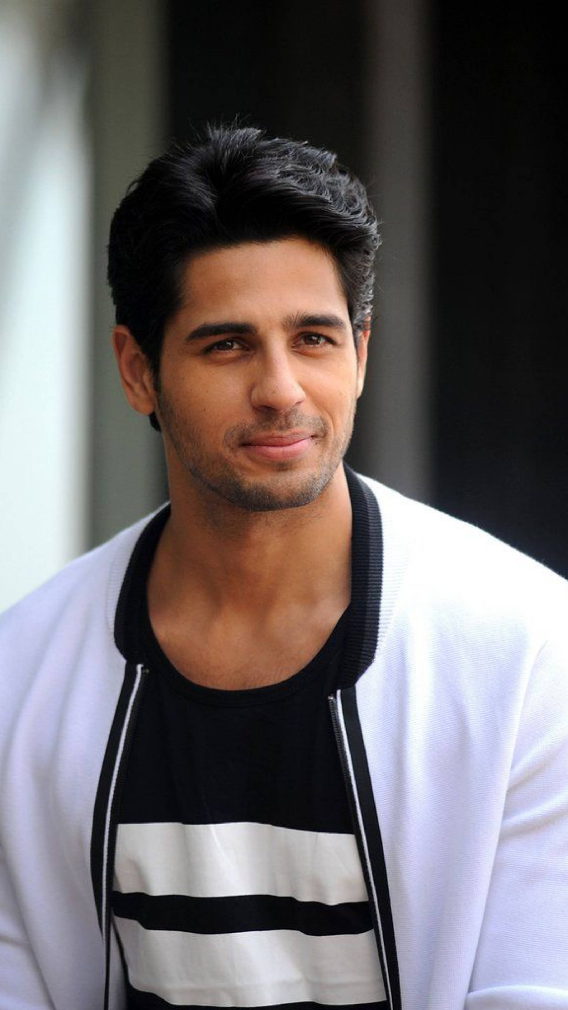 Sidharth Malhotra 桌布 壁紙 — 一位黑髮、留著淺鬍鬚的英俊男士，身穿白色開襟毛衣內搭黑白條紋襯衫，溫柔微笑。
