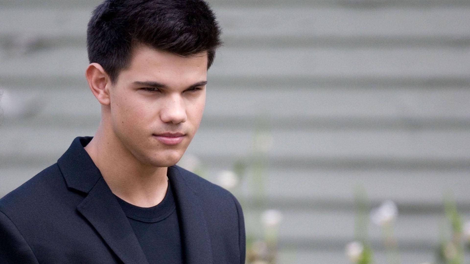 Taylor Lautner 桌布 壁紙 — 一位黑髮、眼神銳利的年輕男子，在模糊的灰色木質背景前，身穿黑色西裝外套內搭黑色T恤。