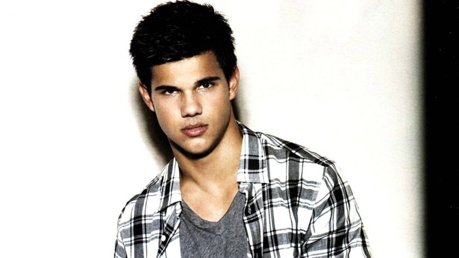 Taylor Lautner 桌布 壁紙 — 一位黑髮、眼神銳利的年輕男子，在純白背景和戲劇性燈光下穿著格紋襯衫。