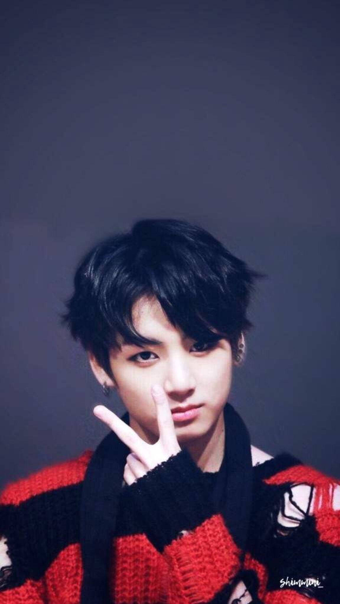 Jungkook Aesthetic 桌布 壁紙 — 一位黑髮、眼神銳利的年輕男子穿著紅黑針織毛衣，在柔和的灰色背景前做出和平手勢。