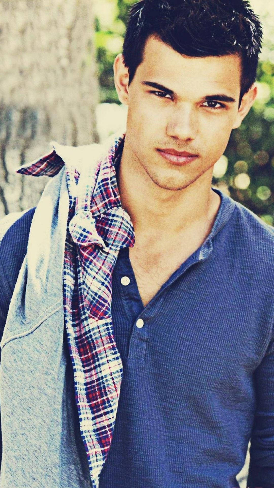 Taylor Lautner 桌布 壁紙 — 一位黑髮、眼神銳利的年輕男子穿著藍色亨利衫，肩上披著一件格紋襯衫，背景是模糊的樹木。