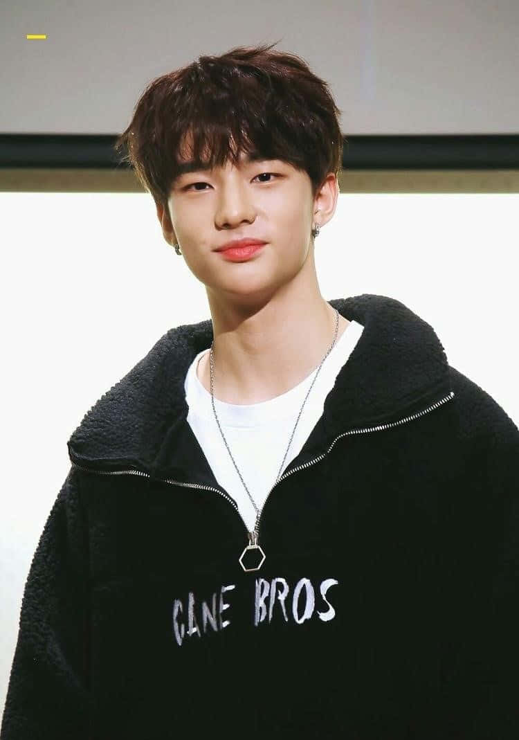 hyunjin 桌布 壁紙 — 一位黑髮、笑容溫和的年輕男子，在純色背景前穿著黑色抓絨連帽衫，內搭白色T恤。