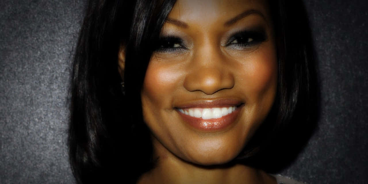 Garcelle Beauvais 桌布 壁紙 — 一位黑髮、笑容燦爛的女士在紋理灰色背景上閃耀著光芒。