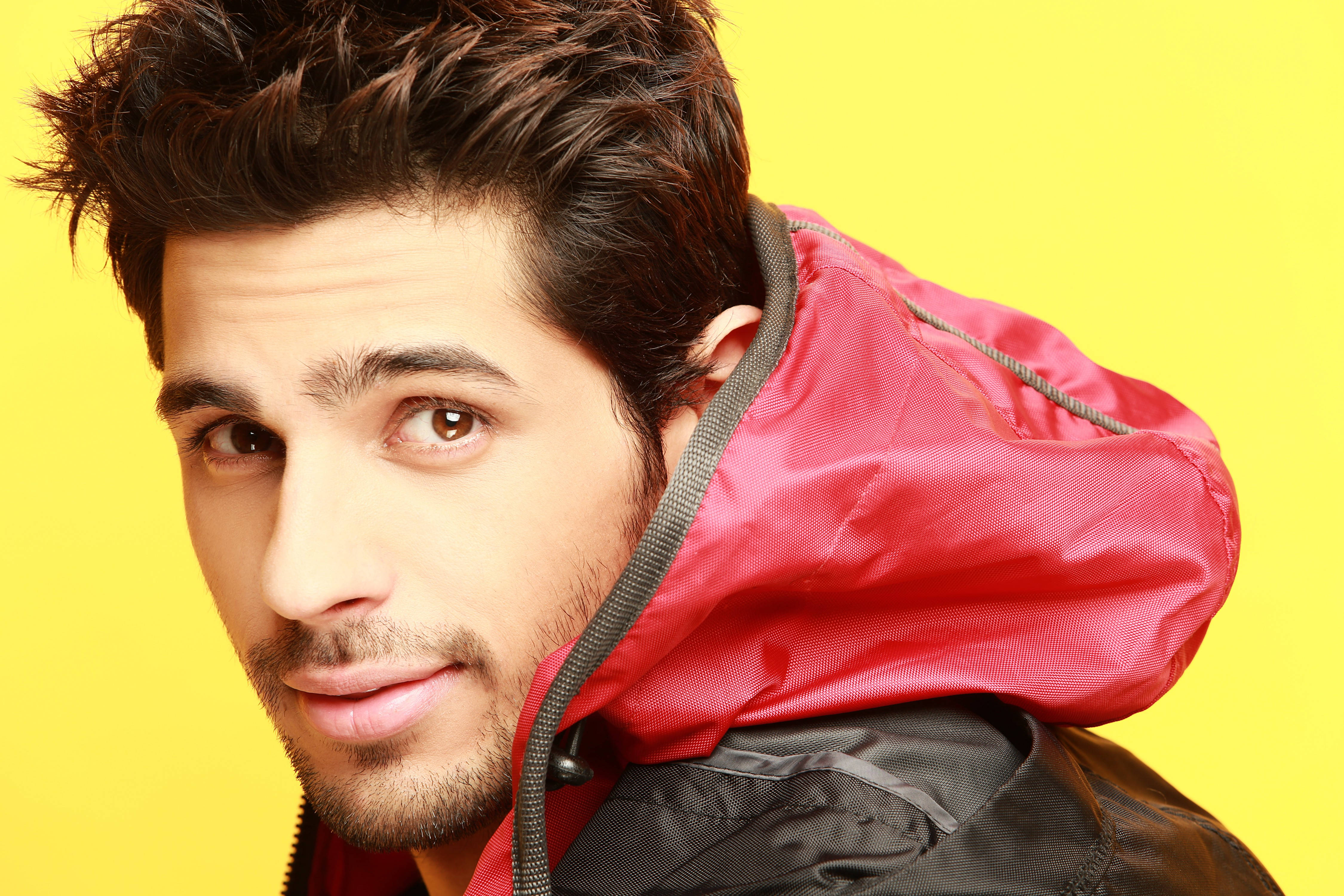 Sidharth Malhotra 桌布 壁紙 — 一位黑髮、身穿紅色連帽外套的年輕男子，在鮮黃色背景前直視鏡頭。